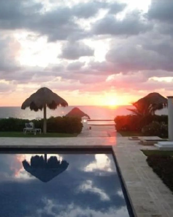 A view of a blue swimming pool and straw cabanas overlooking the sea and sunset.