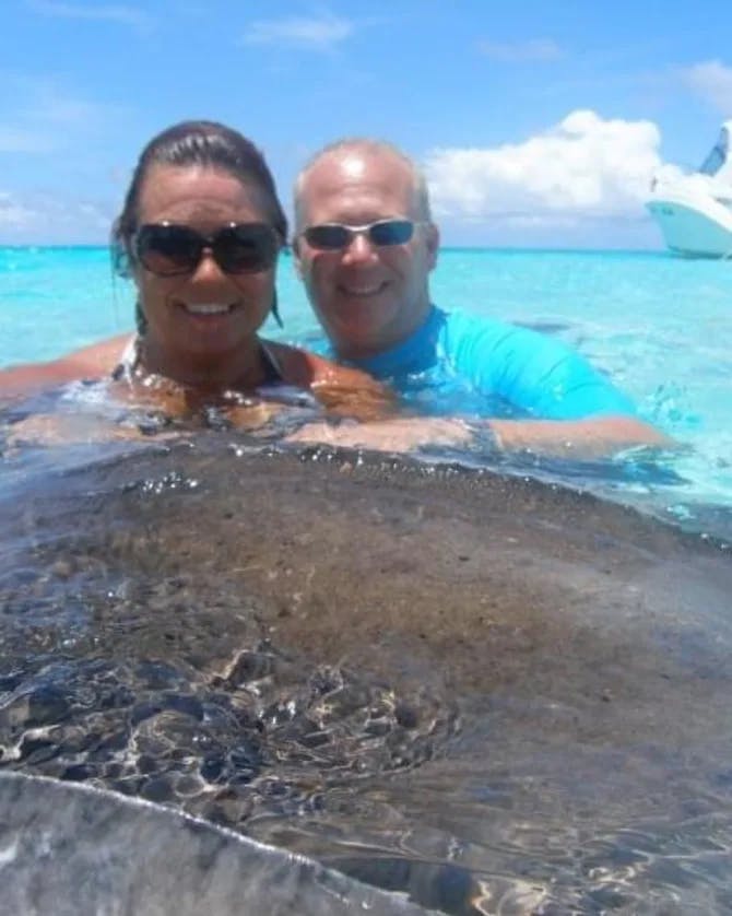 A photo of Robyn and her spouse posing with stingray in the crystal blue water with a boat in the background