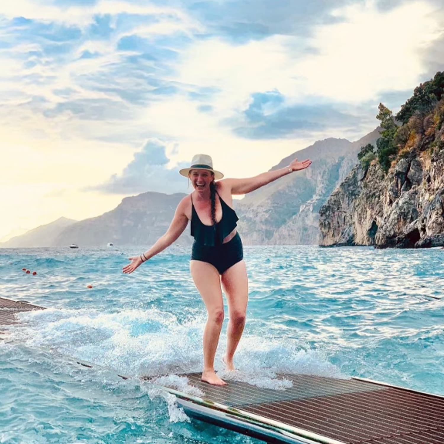 Picture of Heather wearing black swimsuit standing on a platform leading into the ocean with cliffs in the background