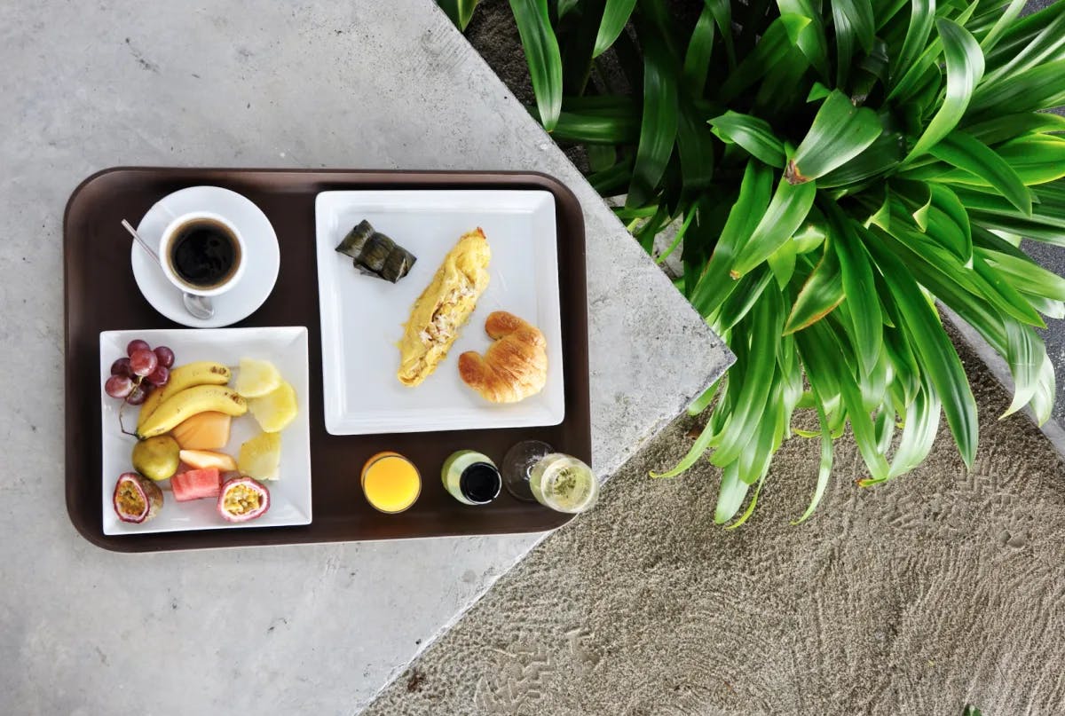 An overhead view of a platter of food with eggs, fruit, coffee and three beverages. There is a green plant and sand next to the platter. 