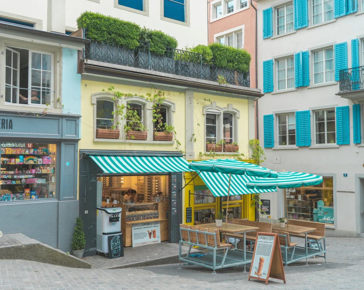 A yellow cafe on a street corner with green and white striped awnings, tables and chairs outside.