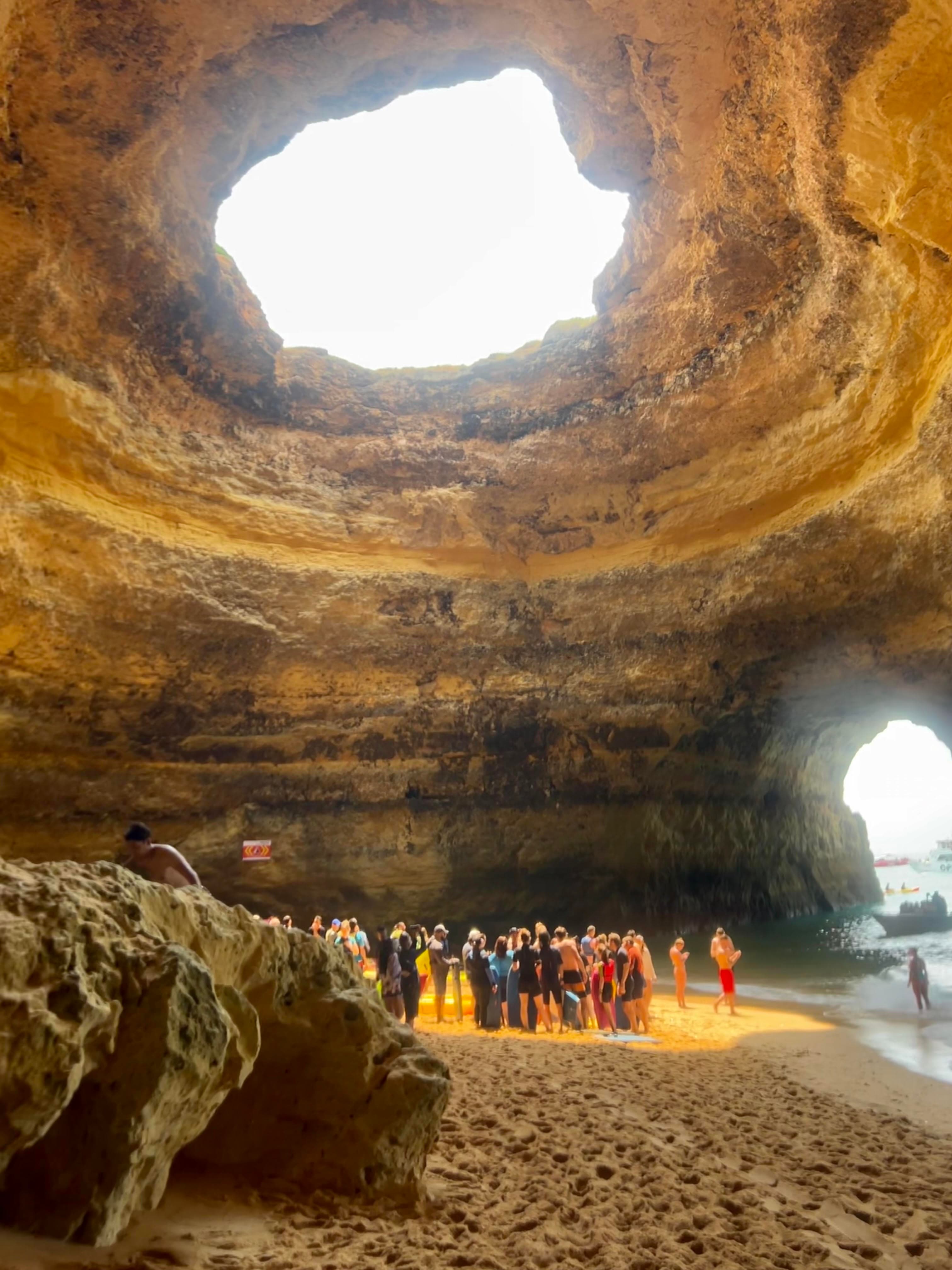 A group of people standing in a beach cave with a hole exposing the sky at the top.