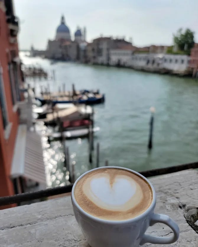 A delicious latte with heart shaped foam perched on a ledge with a canal and beautiful architecture in the distance. 