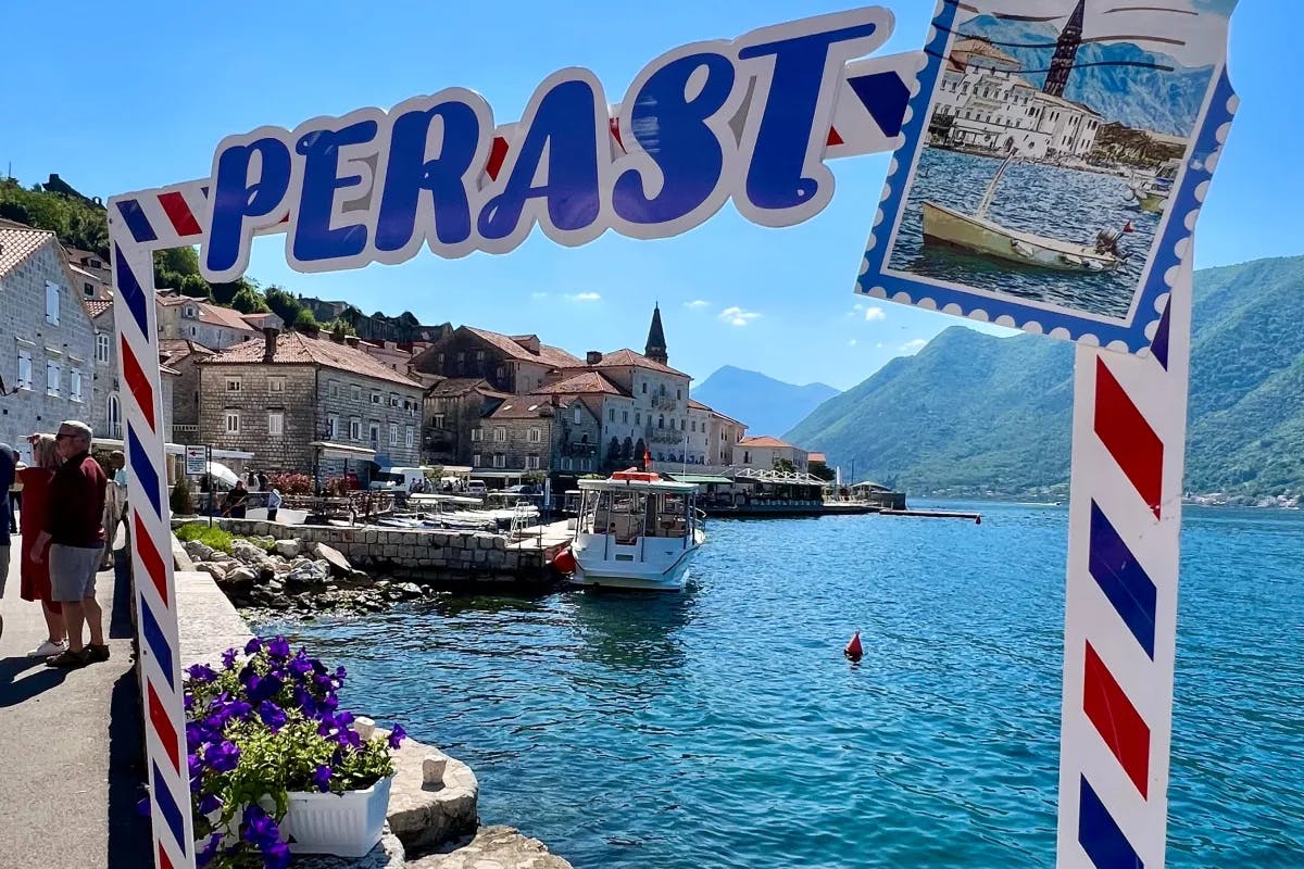 A tourist view of Perast complete with red-roof biuldings, a boat, blue water, green mountains and a red, white and blue frame for photos.