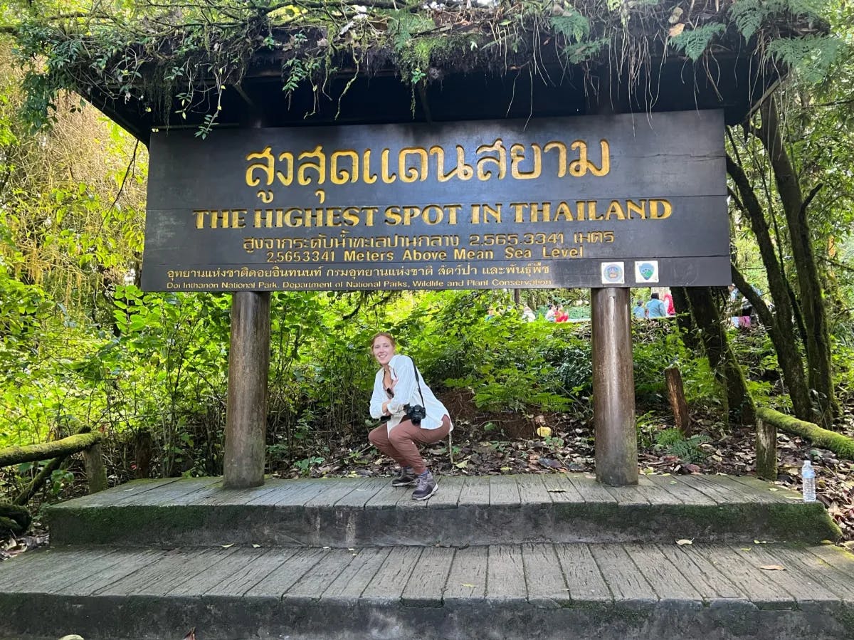 A woman posing in front of a brown sign that reads "The Highest Spot in Thailand" in yellow lettering. 