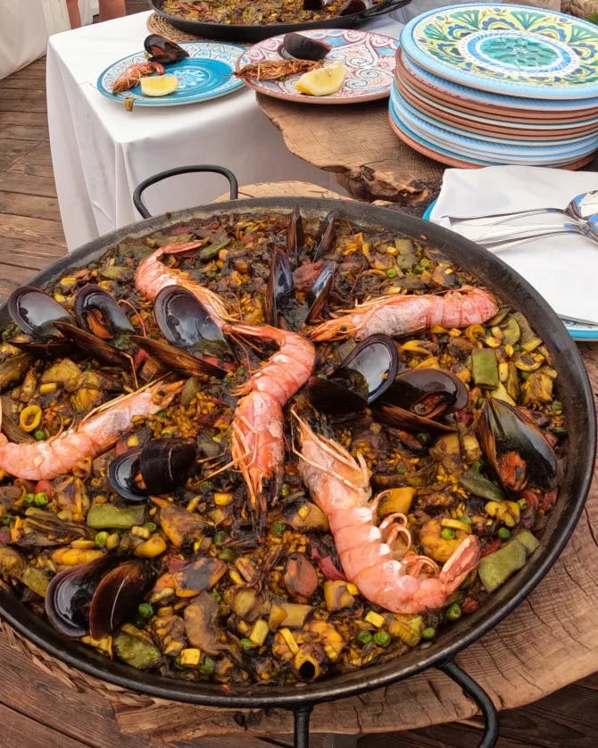 Paella on a wooden table surrounded by colorful plates, spoons and tables in the background.