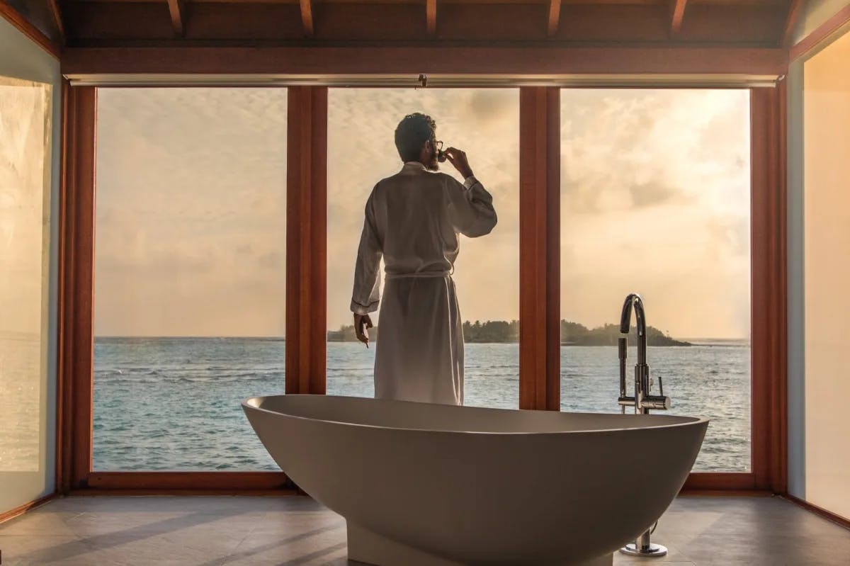 A man in a plush, white bathrobe sips an espresso while admiring the view through floor-to-ceiling windows at a spa in the Maldives