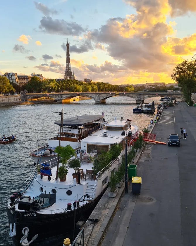 A beautiful view of Paris city and a yacht docked by a road with the Eiffel Tower and a sunset in the background 