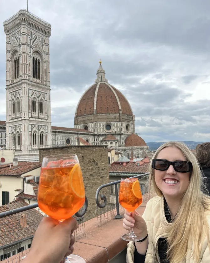 Alyssa and a friend holding an Aperol Spritz while seated in front of a view of the duomo in Florence, Italy