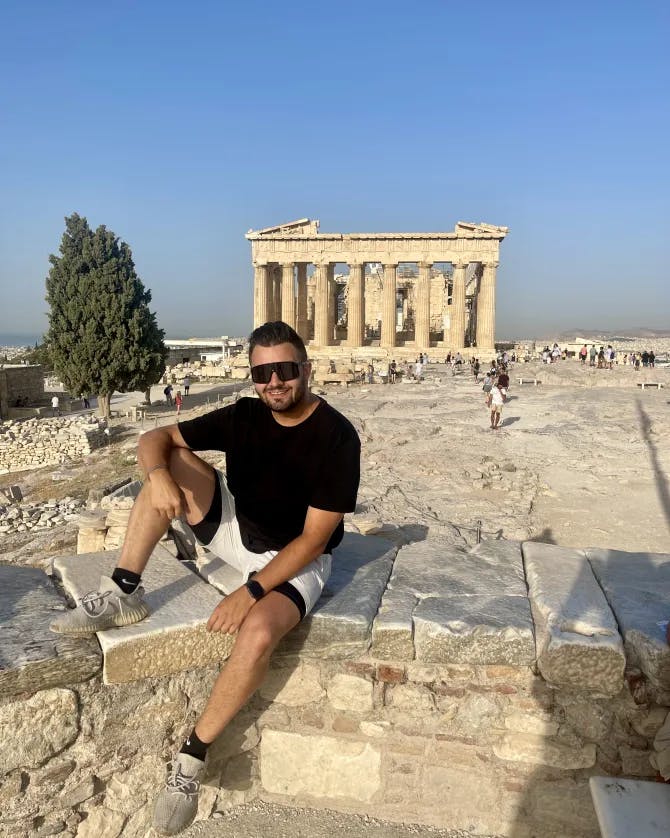 John wearing a black t-shirt and shorts seated on a rock in front of the Parthenon