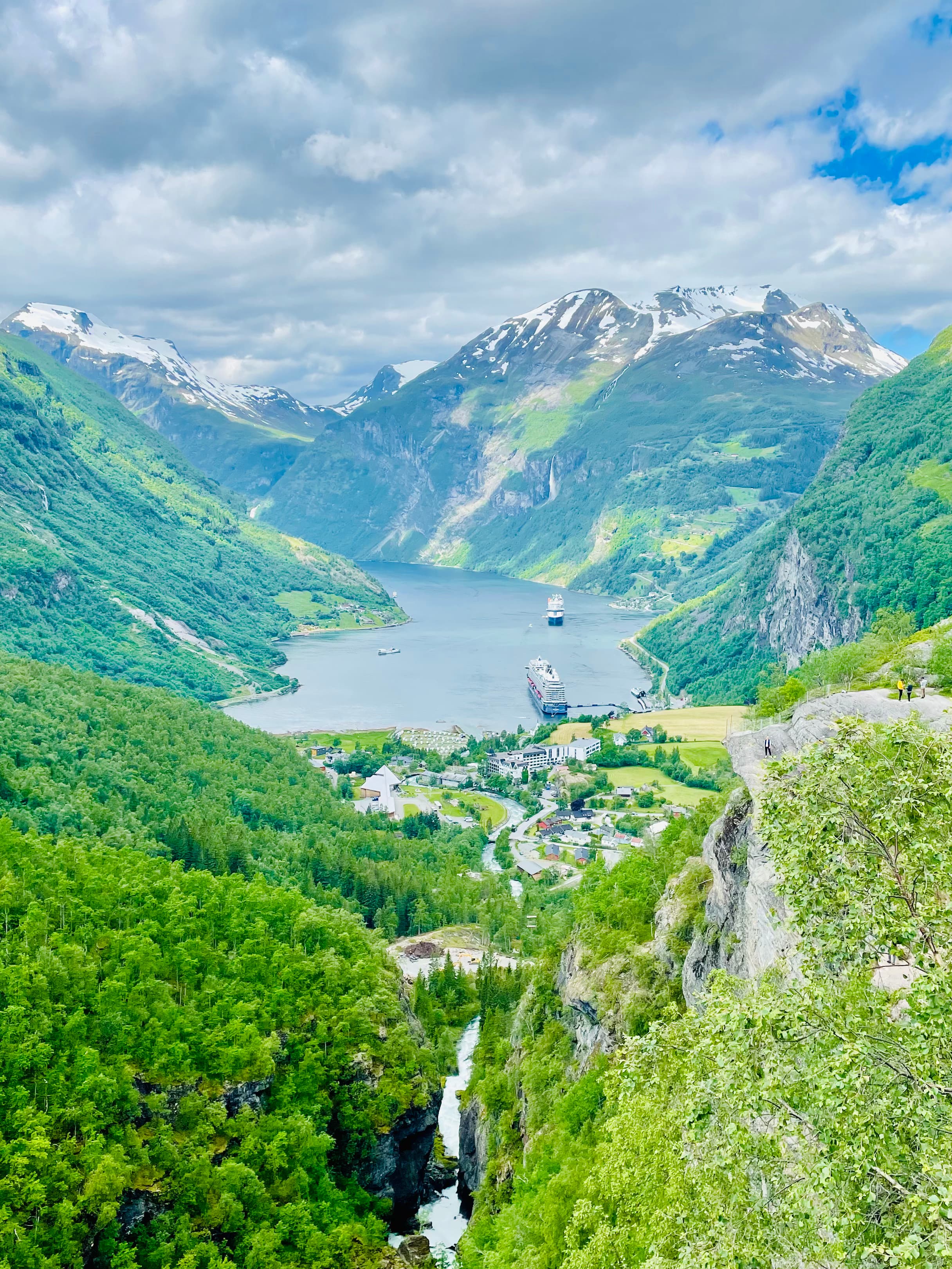 A beautiful view of Geirangerfjord with green trees, a body of water and snowy peaks in the distance.