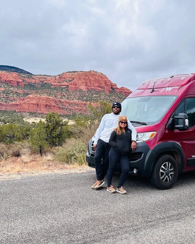 Joanne and partner standing in front of a red van with the desert and a canyon in the background