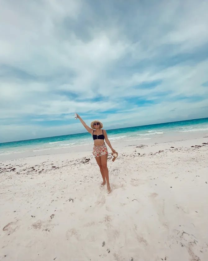 Dylan posing on the beach in a swimsuit with her hand up in the air and the blue water and cloudy sky in the background.