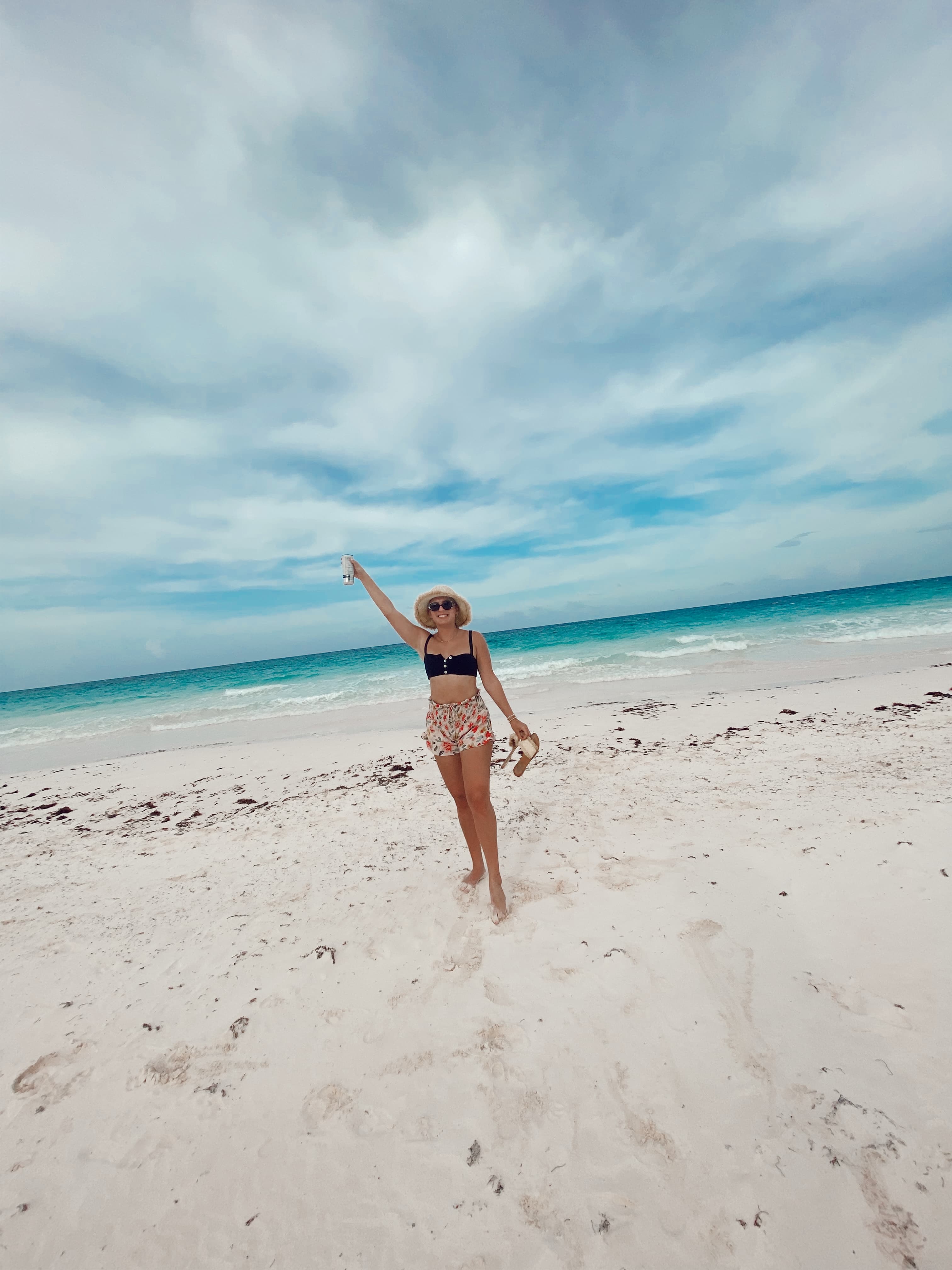 Dylan posing on the beach in a swimsuit with her hand up in the air and the blue water and cloudy sky in the background.