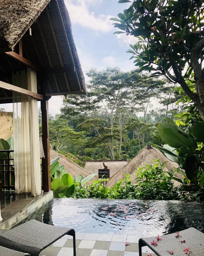 A view from a small sitting deck with a dipping pool that faces other buildings with thatched rooftops and a tropical forest in the distance. 