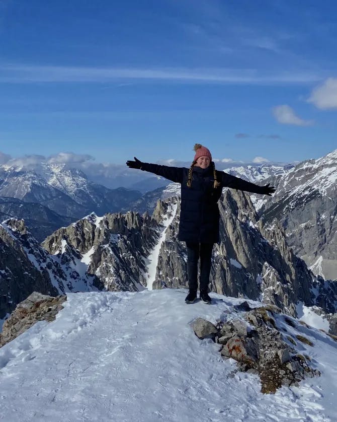 Mackenzie standing on the top of a snowy mountain with her arms stretched out wide and mountain peaks in the surrounding distance 