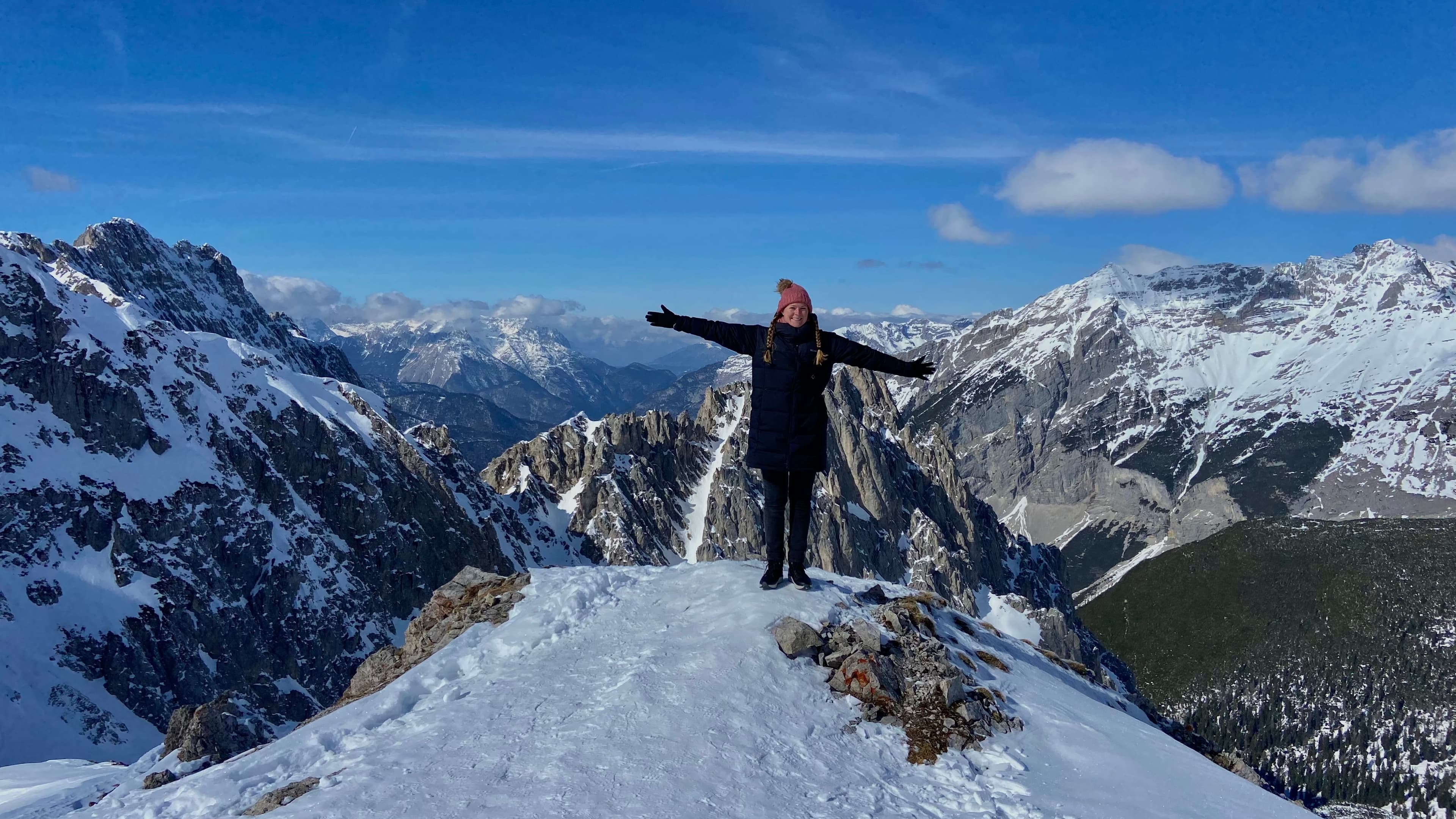 Mackenzie standing on the top of a snowy mountain with her arms stretched out wide and mountain peaks in the surrounding distance