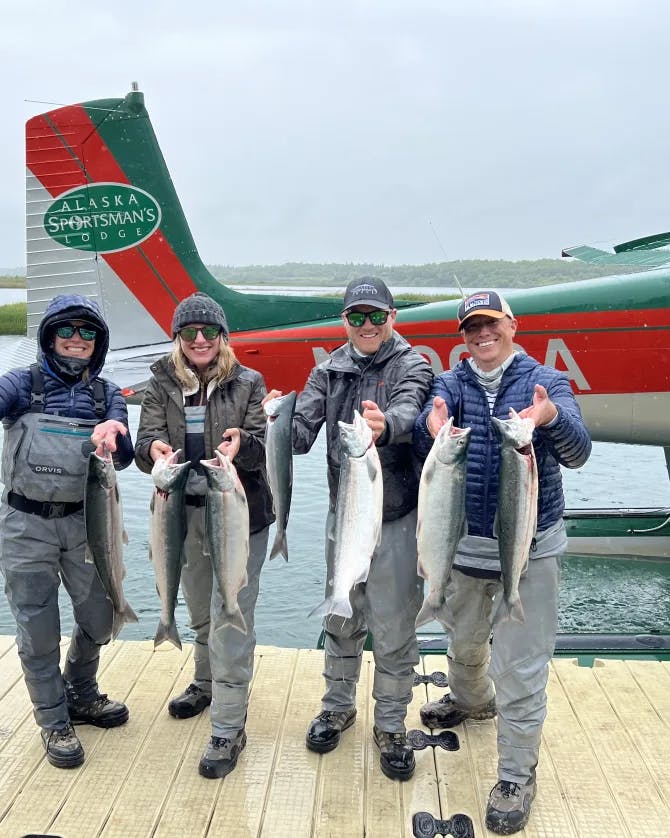 Four people posing with fish while standing on a wooden dock in front of a water plane and water 