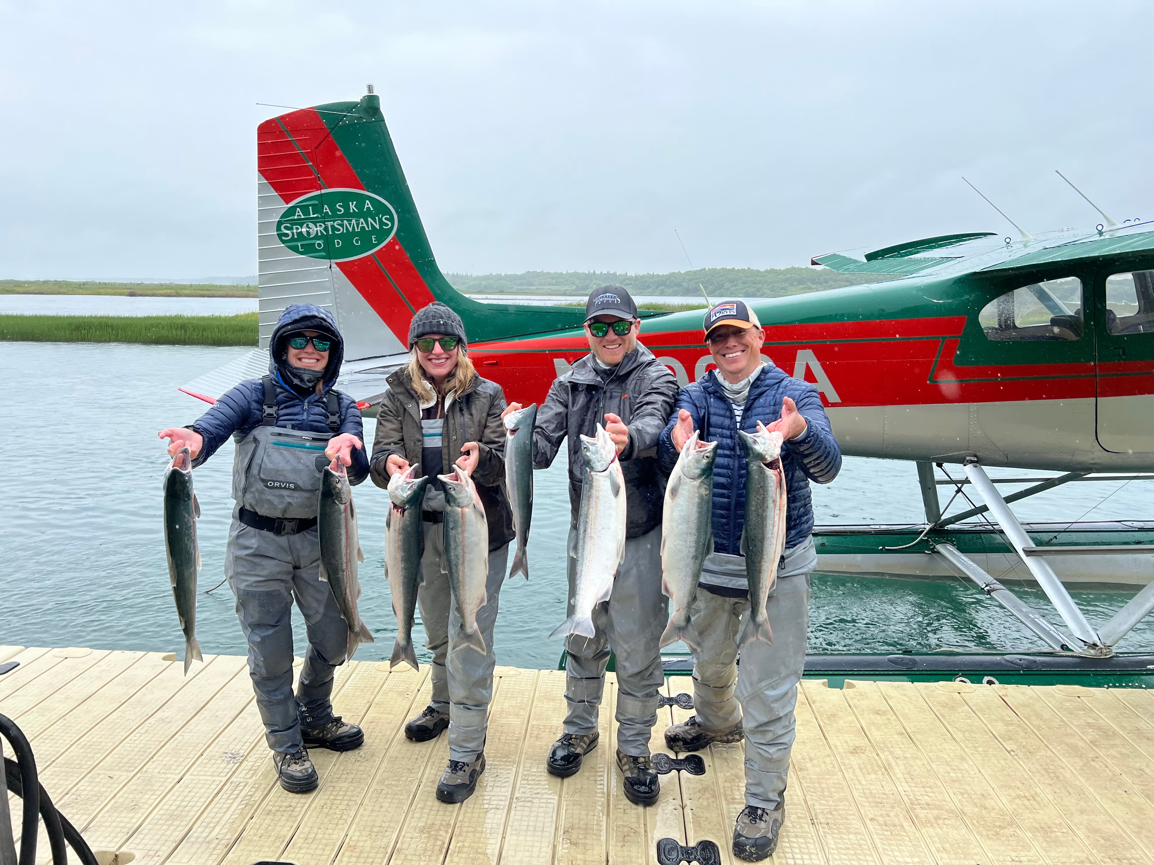 Four people posing with fish while standing on a wooden dock in front of a water plane and water