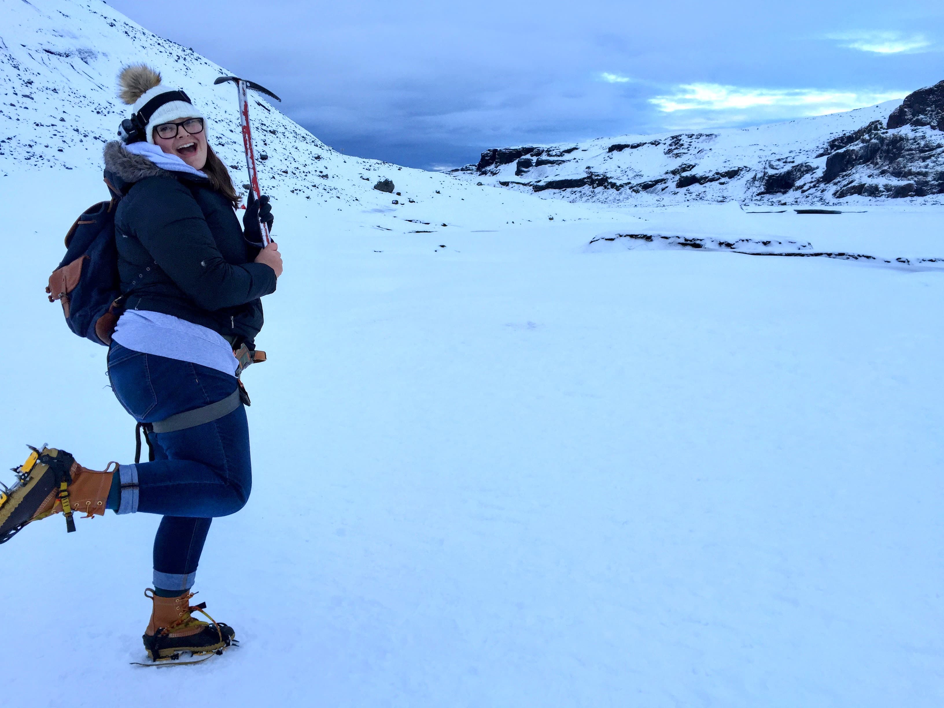 Jamie wearing winter gear posing outside with a pick axe with snowy mountains in the background