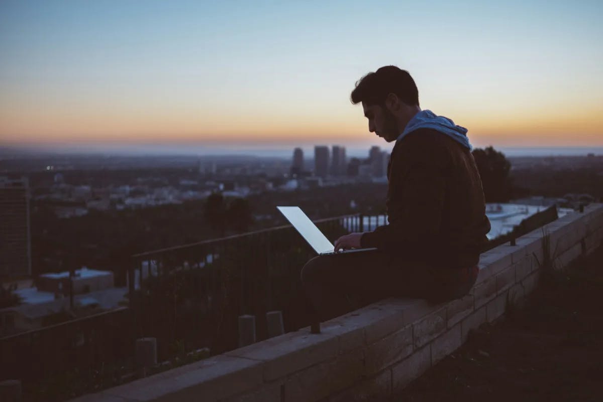 At dusk, a man in trendy attire works on a laptop from a rooftop with an urban skyline in the distance