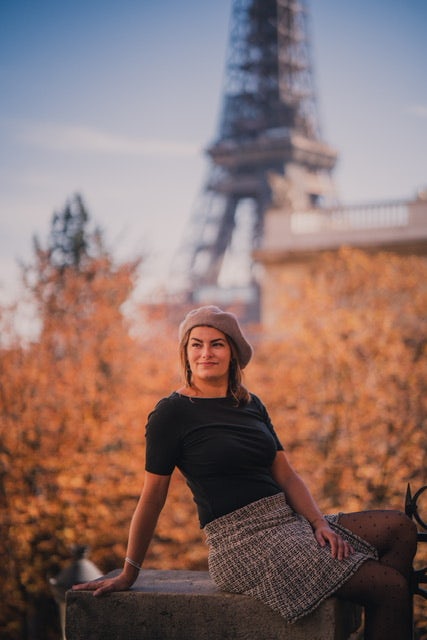 travel advisor Jovana Pellerito sits in front of the Eiffel Tower and autumn trees wearing a black shirt and baret