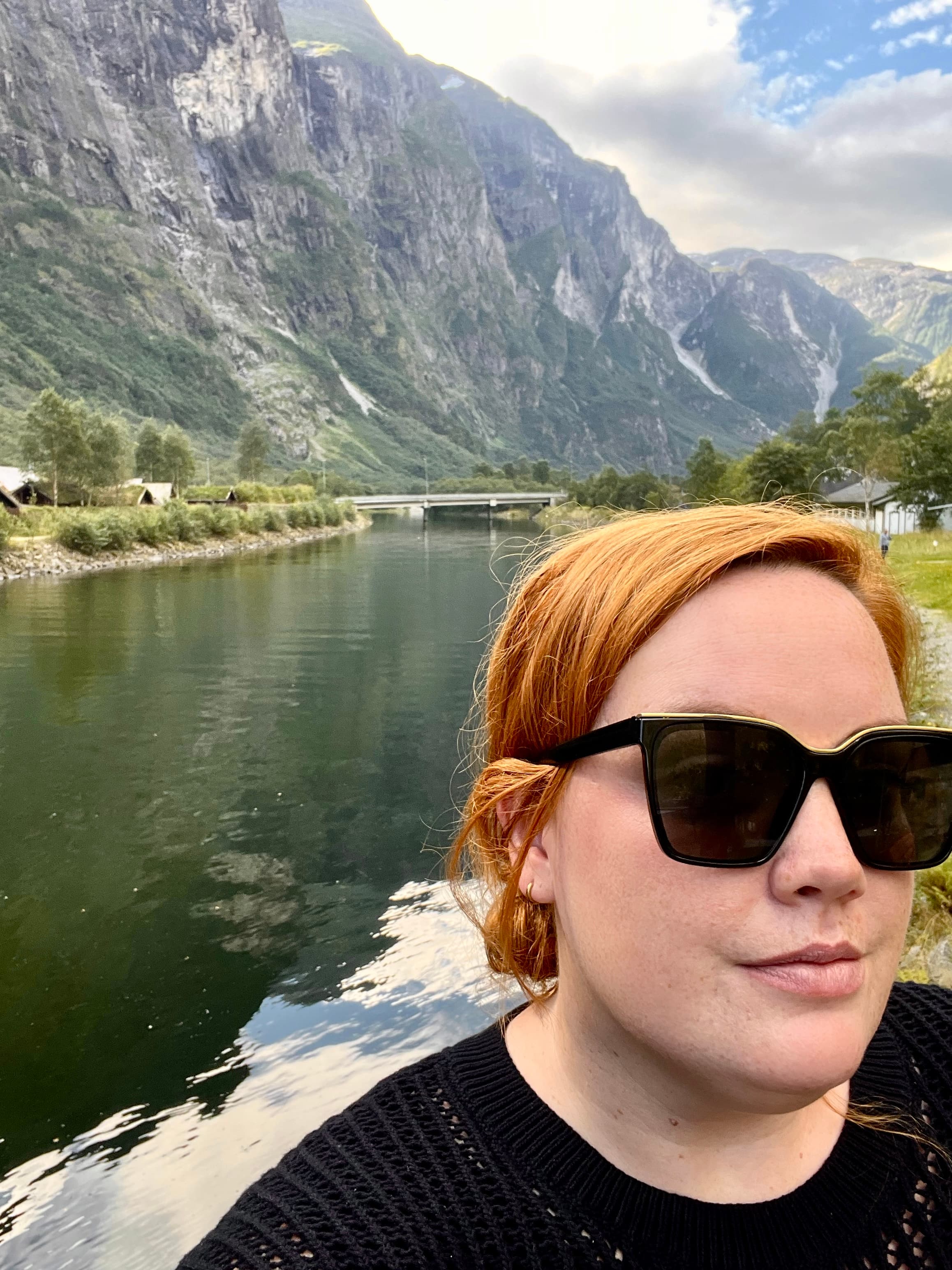 Emyli wearing a black top and sunglasses posing for a selfie in front of a lake and mountains
