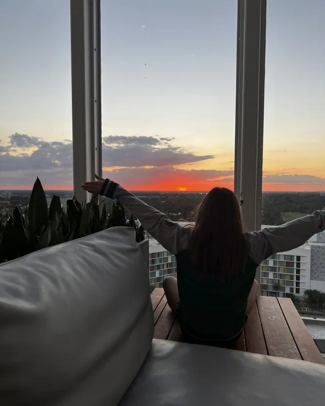 Picture of Amanda watching sunrise from a bedroom while sitting on a wooden floor with her arms spread open 