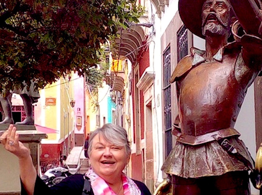 Travel Advisor Mary Manix stands next to a bronze statue on an old cit street with color buildings behind her 