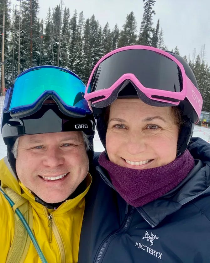 Kate and partner posing for a selfie while wearing ski gear in front of pine trees and a snowy ski hill