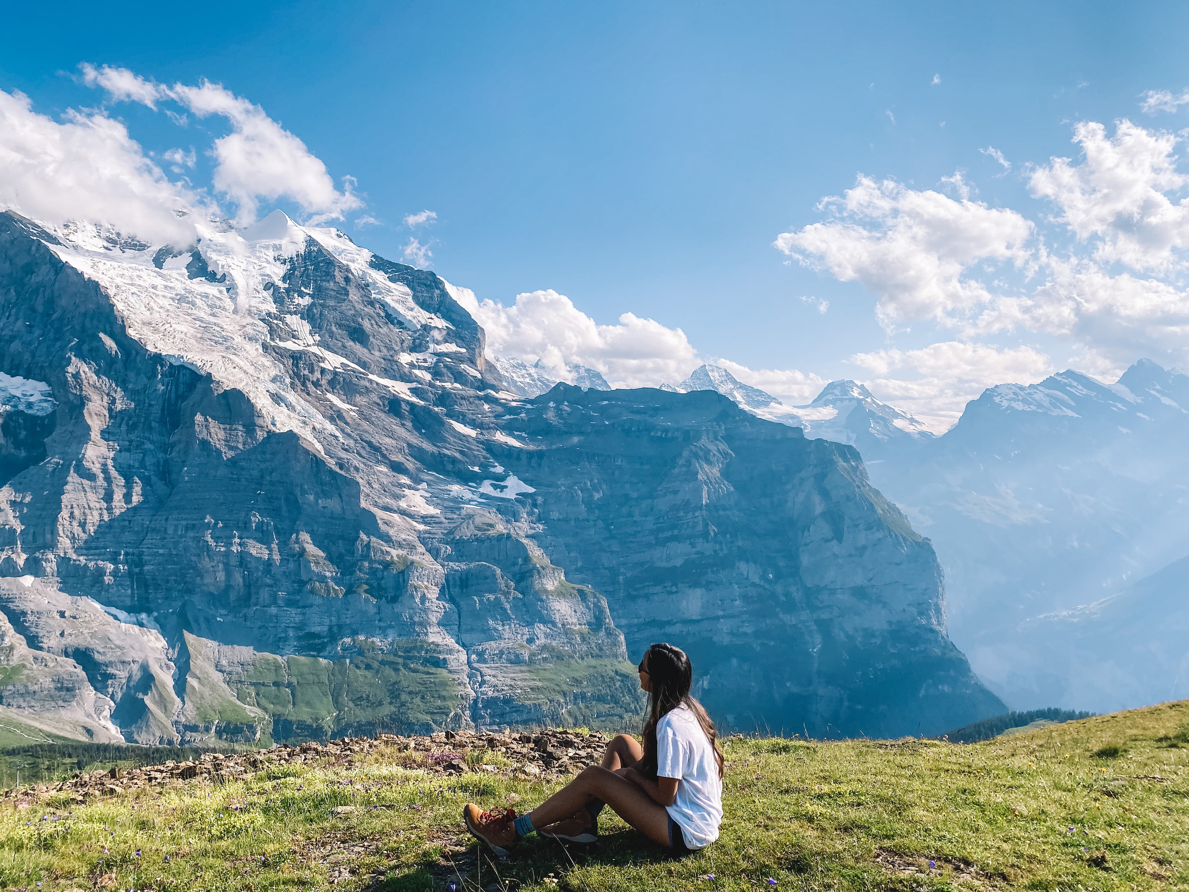 Travel advisor sitting on the grass near a cliff overlooking a mountain range.