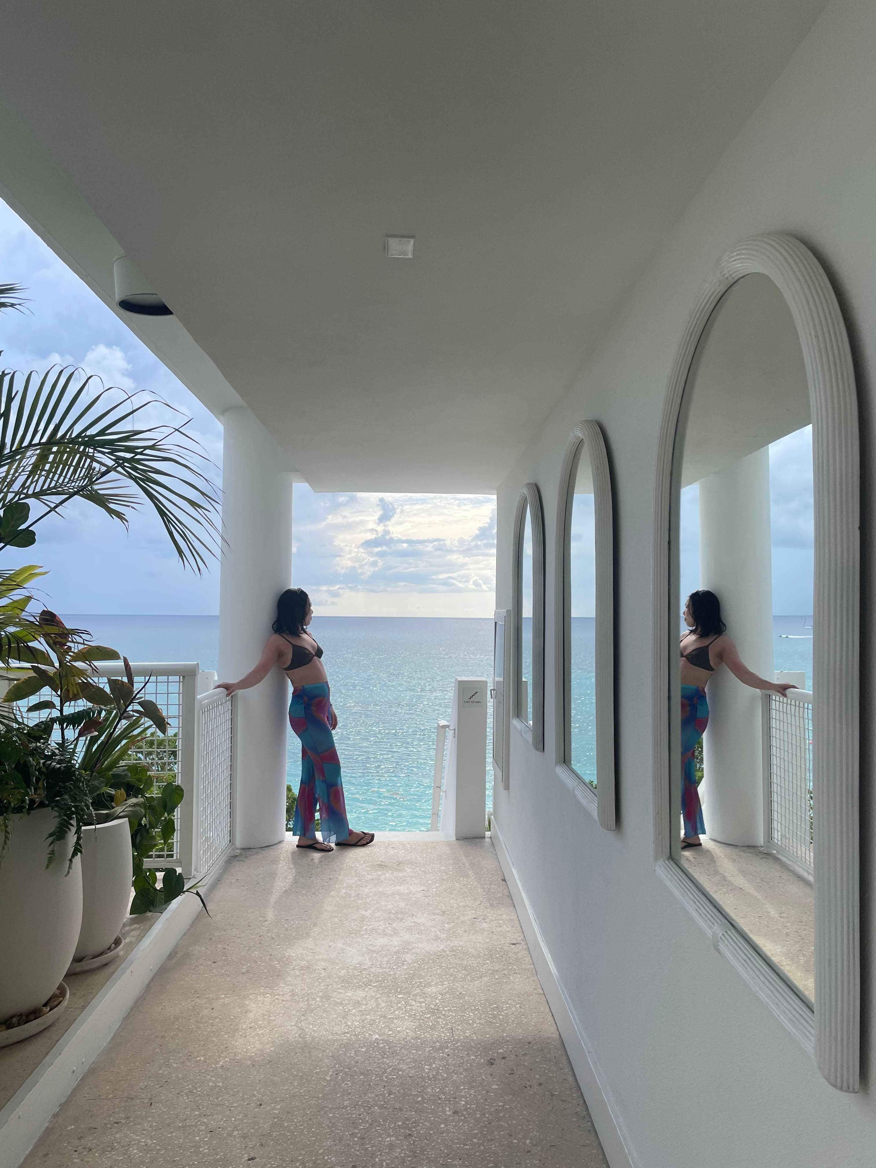 Kimberly posing at the end of an outdoor hallway decorated with white arched mirrors, white walls and green plants in pots that overlook the blue water.