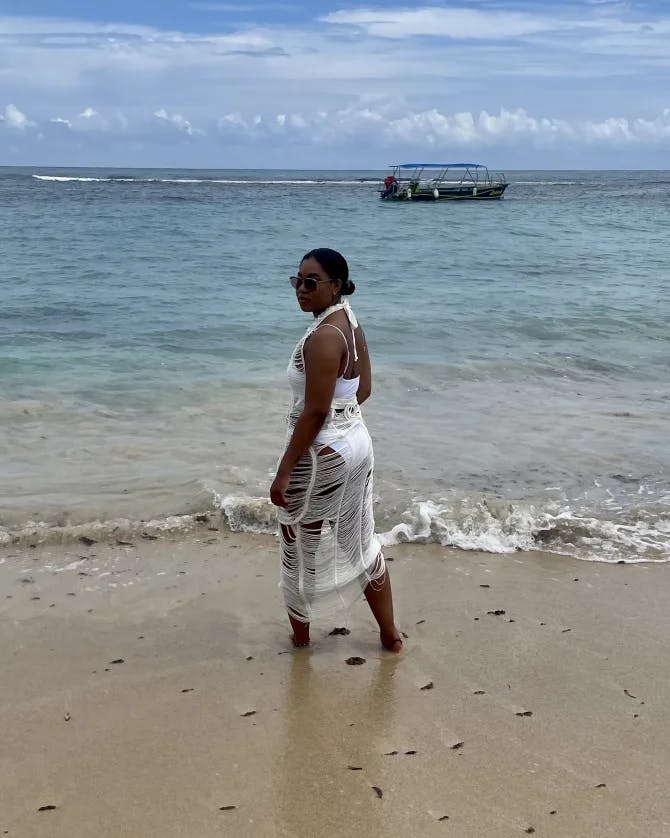 Tyler posing in a white dress on a sandy beach with blue water and a boat in the background.