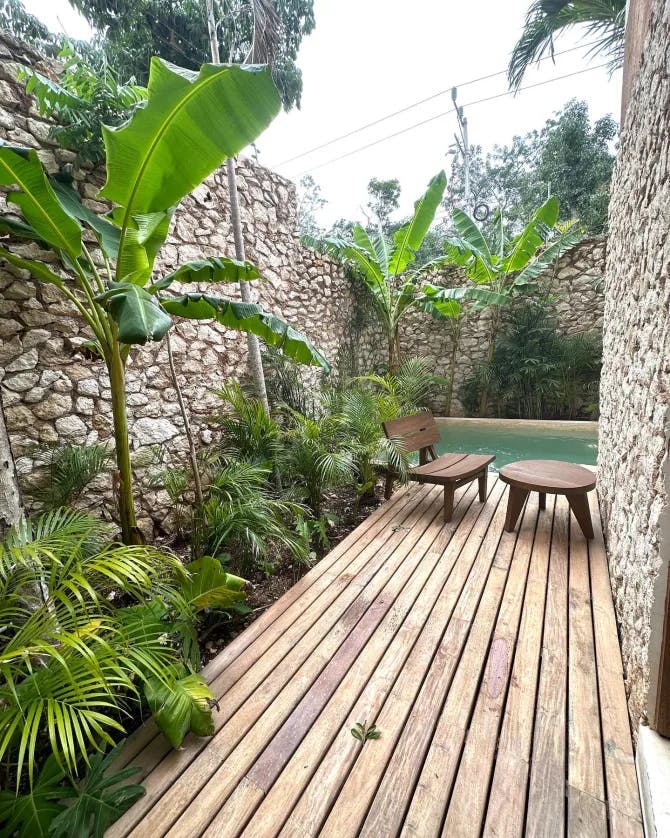 A wooden deck next to a bench, table, stone wall, palm trees and water