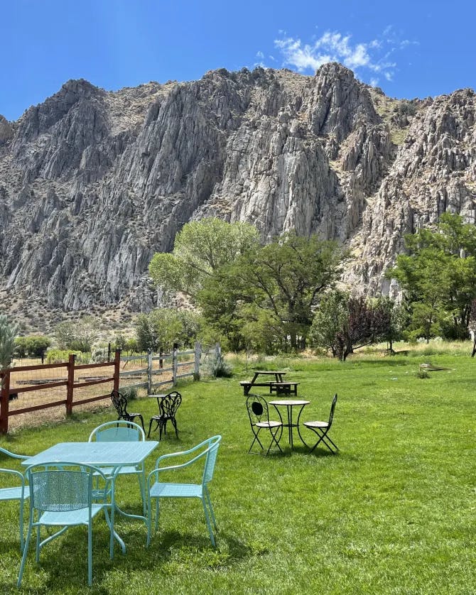 A set of blue chairs and a matching table on green grass next to a fence with a rocky canyon and other tables and chairs in the background 
