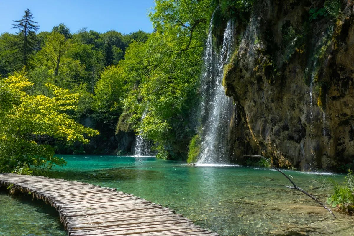 A wooden pathway surrounded by crystal green water, a stone wall, waterfall and bright green trees.
