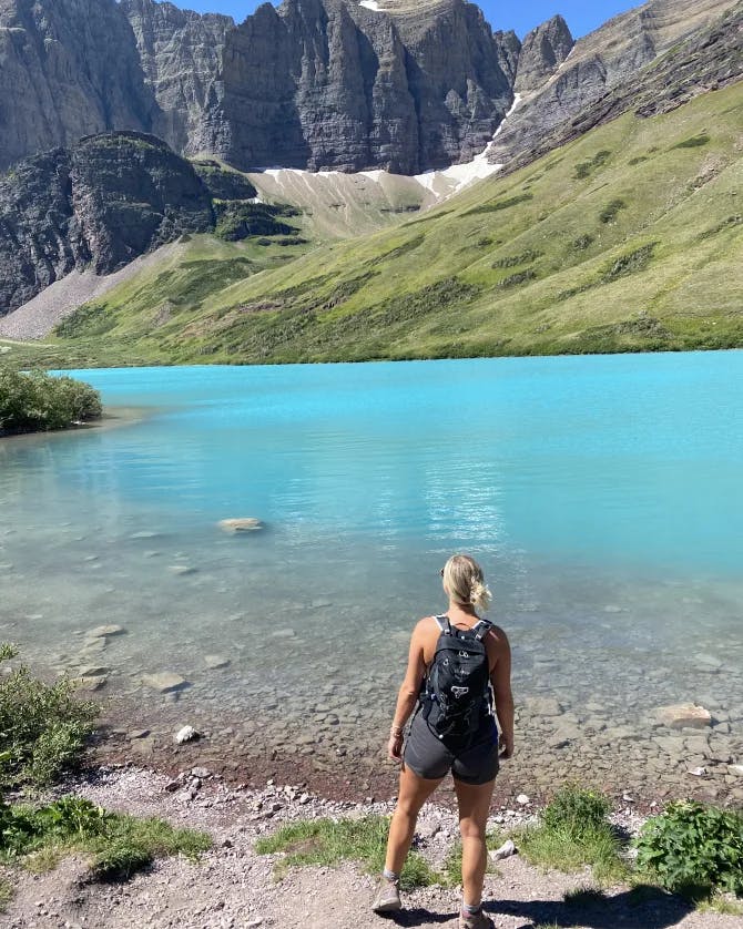 Samantha wearing athletic gear and standing on a dirt path that leads to a bright blue lake surrounded by green and rocky mountains
