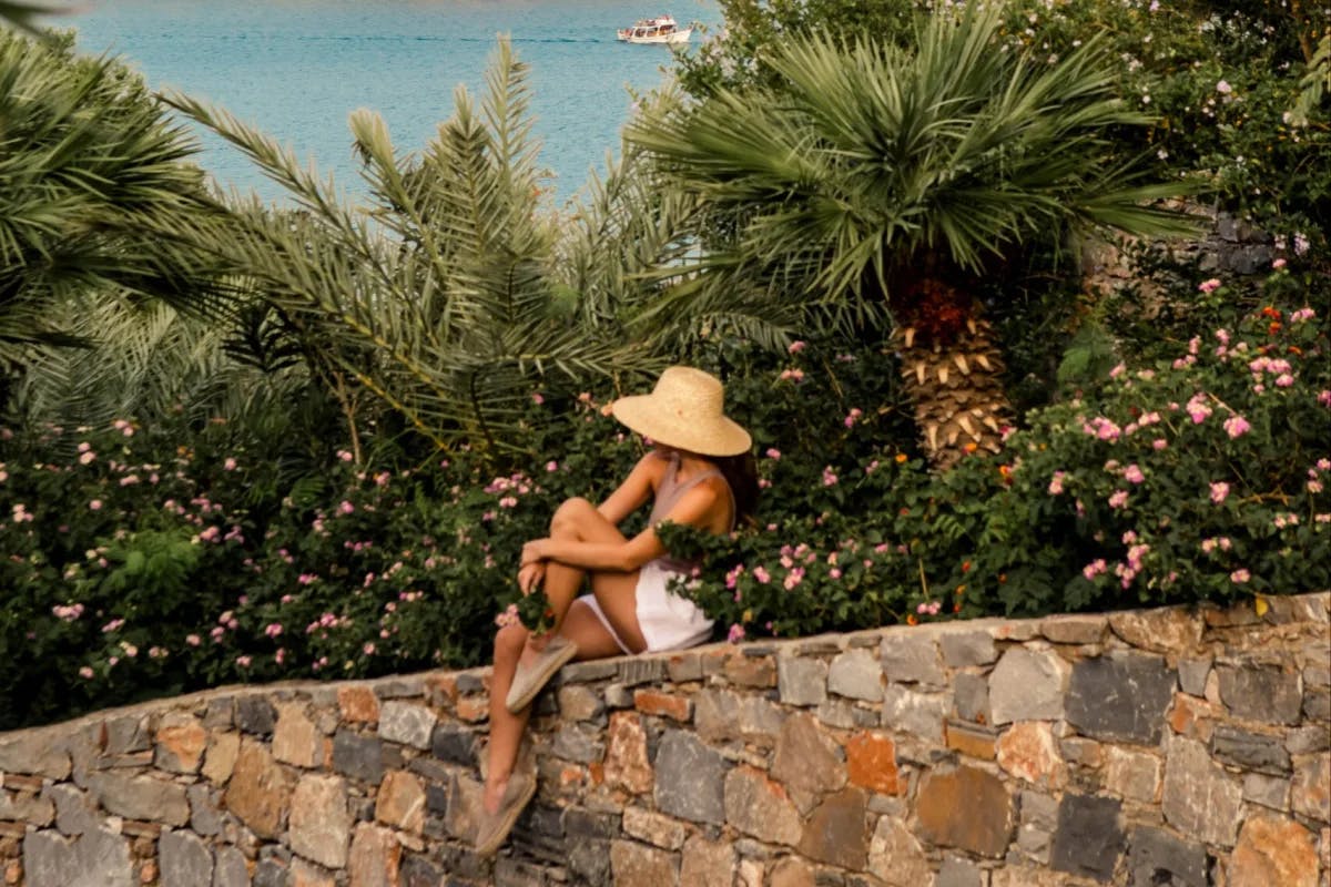 a woman in a large sunhat sits on a stone wall overlooking the sea