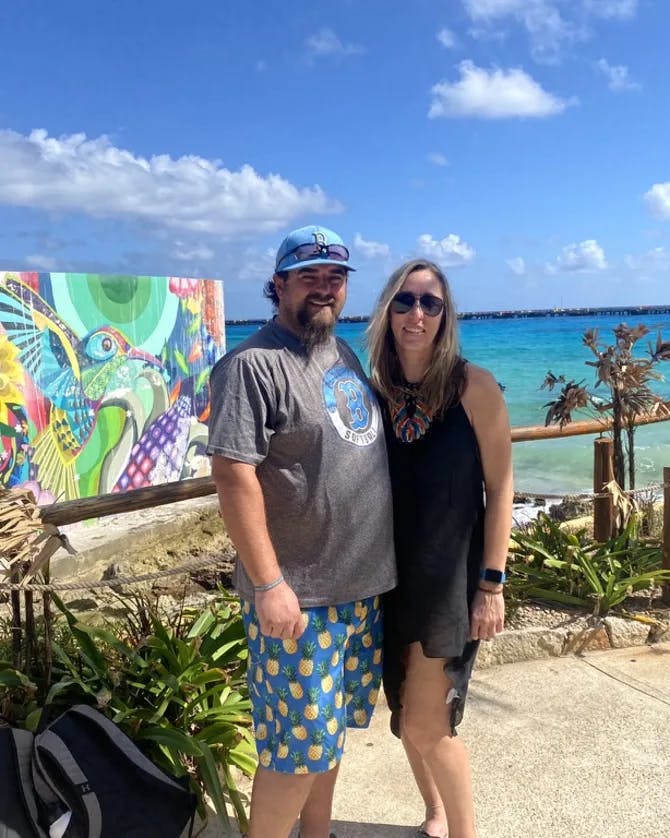 Picture of Amanda in a black dress with her husband in front of a colorful mural and sea view