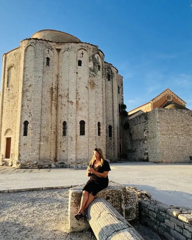 Karyn sitting in Zadar with an ancient town setting behind her and clear skies. 