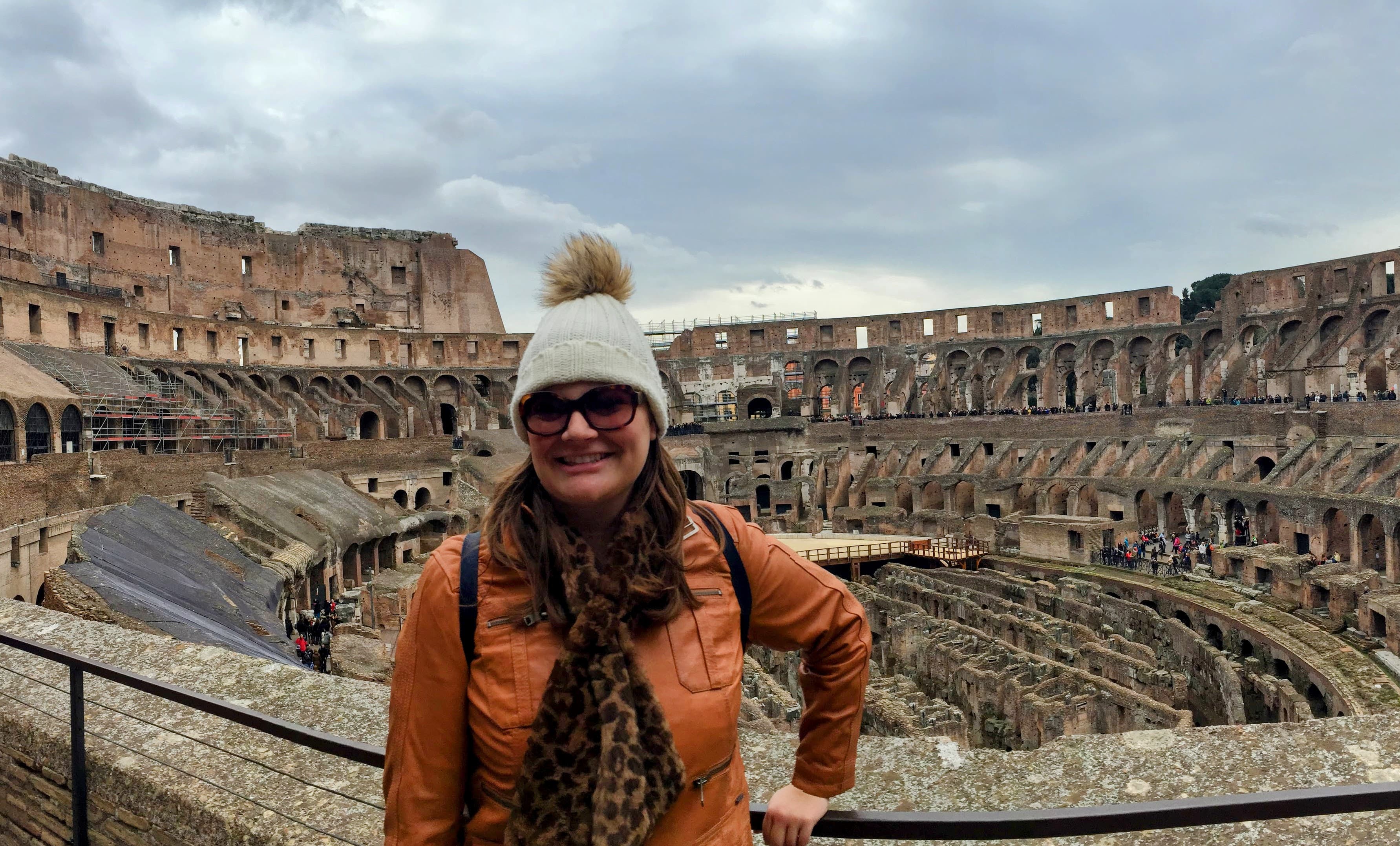 Jamie wearing an orange jacket and winter hat while posing in front of the colosseum in Rome, Italy