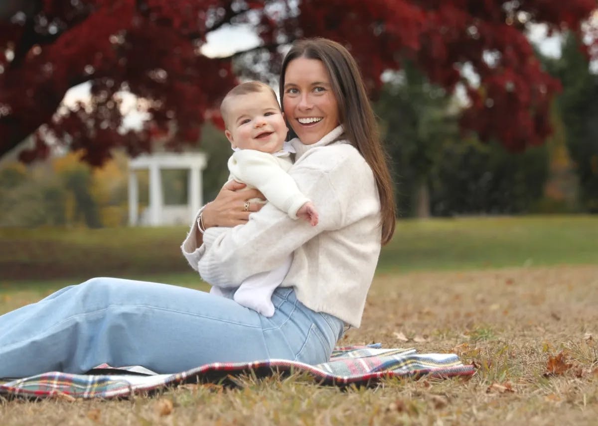 a woman in a white sweater holds a baby on a picnic blanket
