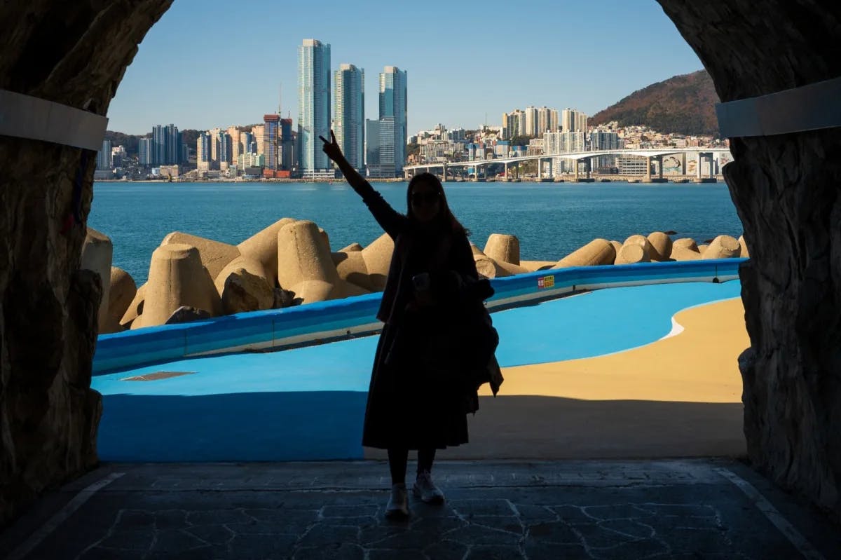 A young woman stands at the end of a tunnel, making a peace sign. Directly behind her is a small bay leading to the skyline of Busan, South Korea