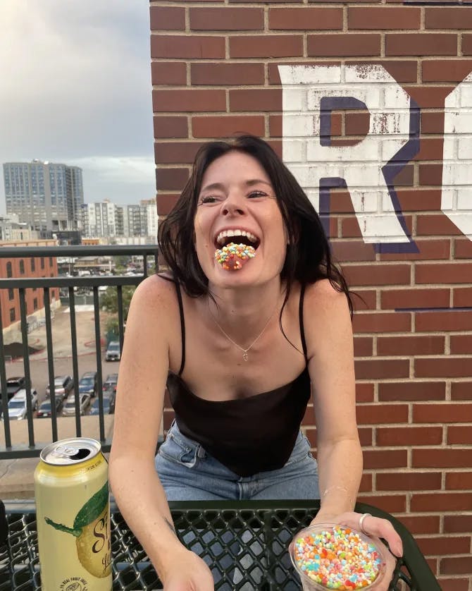 Picture of Bridget eating and laughing in front of a brick wall with a cityscape in the background