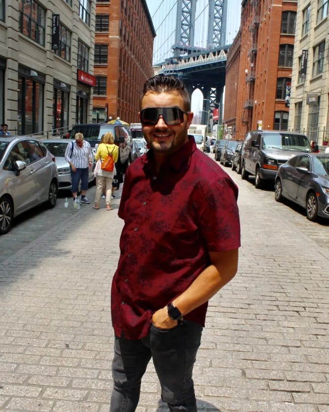John wearing a red top and black pants standing on a brick road in front of the Manhattan Bridge with buildings and parked cars in the background