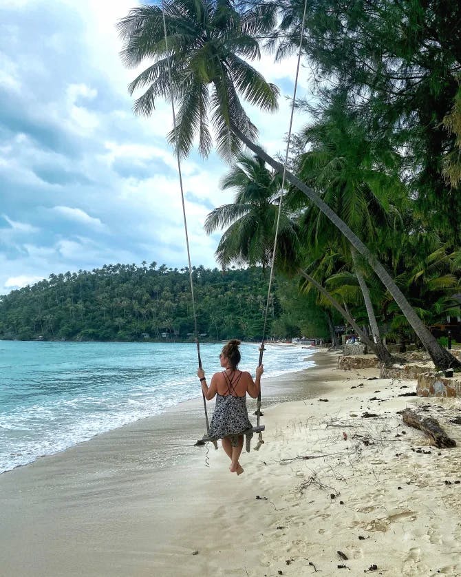 Picture of Marisa on a swing near the ocean with a lovely shoreline with foliage and palm trees. 