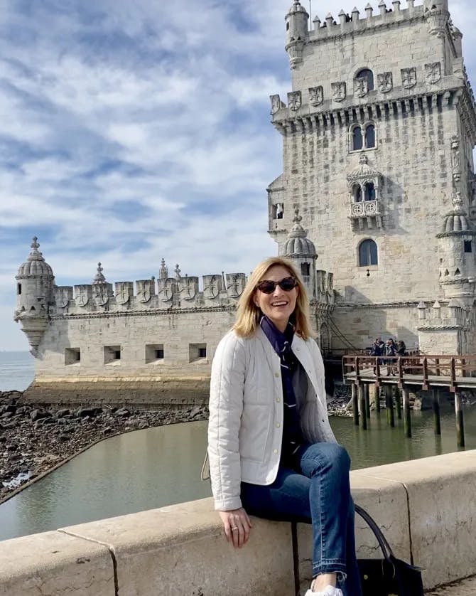 An image of Kristy wearing a white coat and sitting on a stone ledge in front of the Belém Tower