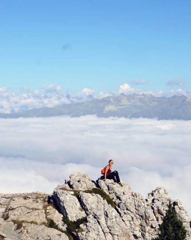 A person sitting at the peak of a rocky mountain surrounded by clouds and blue sky