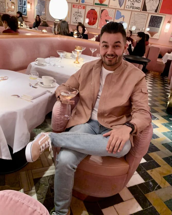 Picture of John sitting in a pink chair with a wine glass in hand at a luxurious restaurant with art and tables in the background
