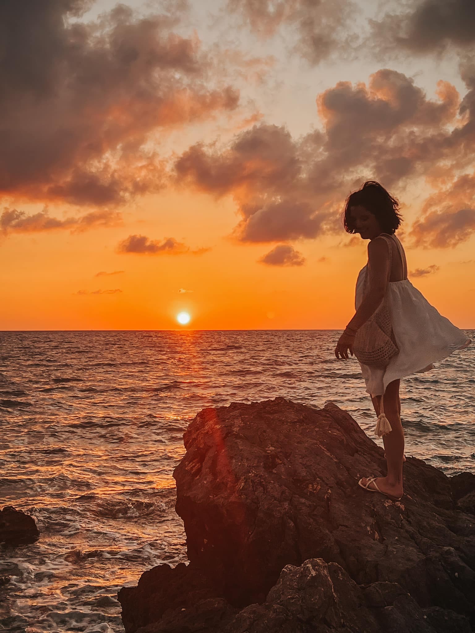 A woman in a white dress standing on a rock overlooking the ocean at sunset